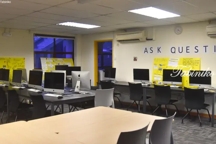 Modern computer lab with Apple iMacs, desks, and chairs, featuring an &lsquo;ASK QUESTIONS&rsquo; wall sign.
