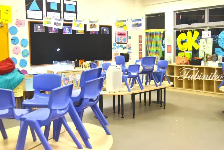 Blue plastic chairs stacked on tables in a colorful classroom with educational posters and a chalkboard.