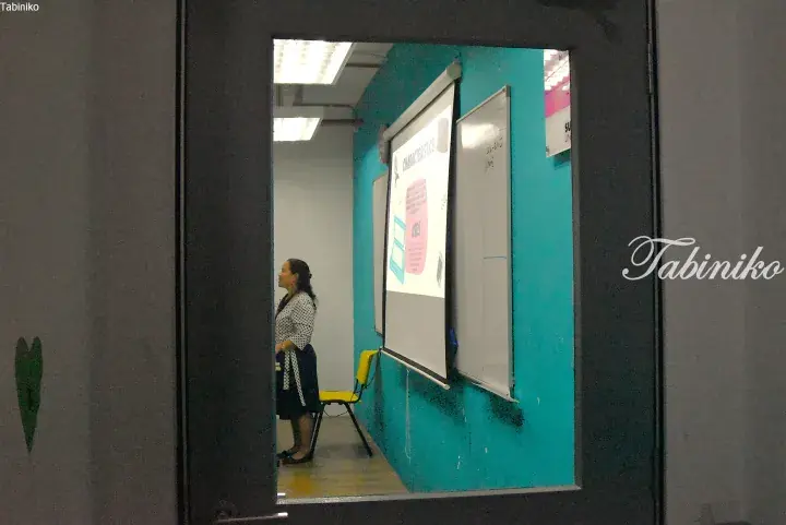 Woman teaching in a classroom with a projector screen showing characteristics and uses, viewed through a glass door.