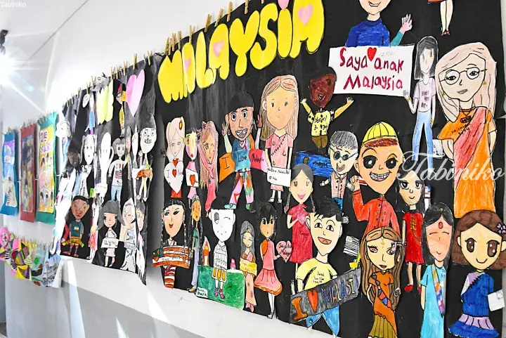 Children&rsquo;s drawings of diverse Malaysian people displayed on a blackboard with &lsquo;Malaysia&rsquo; and &lsquo;Saya anak Malaysia&rsquo; written on it.