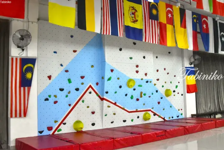 Indoor climbing wall with colorful holds and red mats, decorated with international flags in a Malaysian venue.