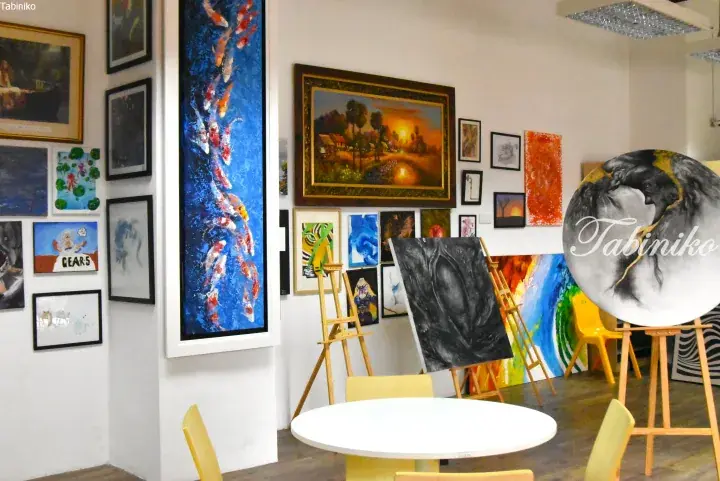 Gallery space displaying diverse paintings on white walls, including a large koi fish artwork, with wooden easels and a round table in the foreground.