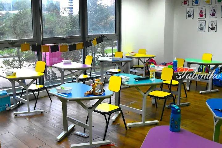 Colorful classroom with child-sized tables and chairs near large windows overlooking greenery.