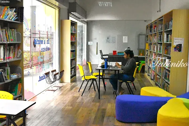 Woman working at a table in a bright library with bookshelves and colorful seating.
