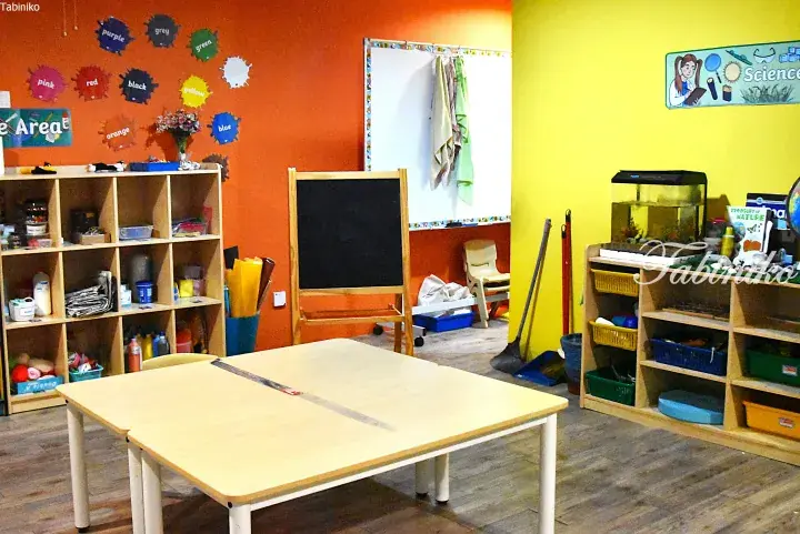 Colorful classroom with orange and yellow walls, wooden shelves, a table, and educational decorations.