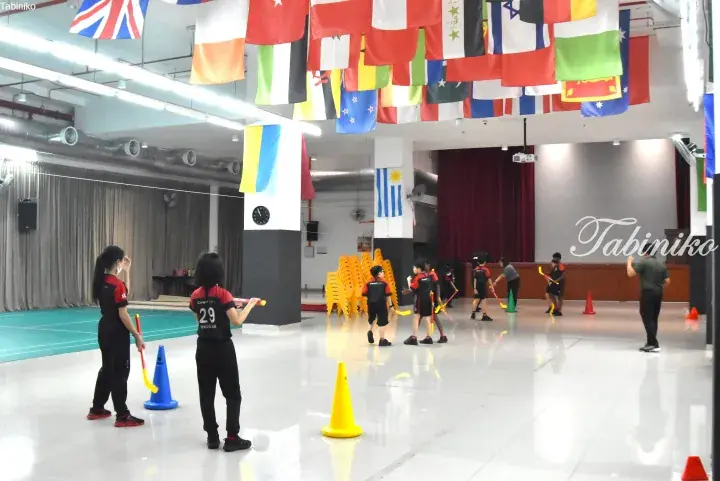 Children practice indoor hockey with sticks and cones under international flags at a Malaysia school hall.