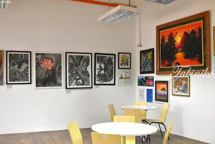 Gallery space with framed artwork on white walls and yellow chairs around white tables.