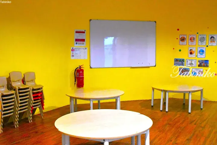 Bright yellow classroom with round tables, stacked chairs, whiteboard, and educational posters.