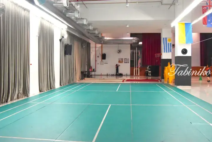 Indoor sports hall with green badminton court, stage, and international flags at a Malaysia school or community center.