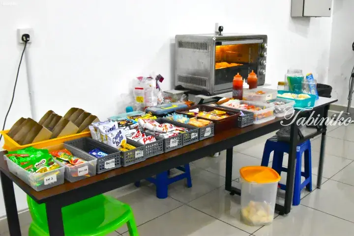 A table displaying various snacks and condiments with a toaster oven, likely a self-service pantry in a Malaysian property.