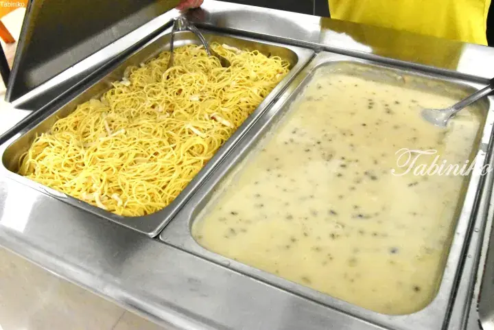 Buffet serving station featuring a tray of yellow noodles and a creamy soup with herbs in stainless steel containers.