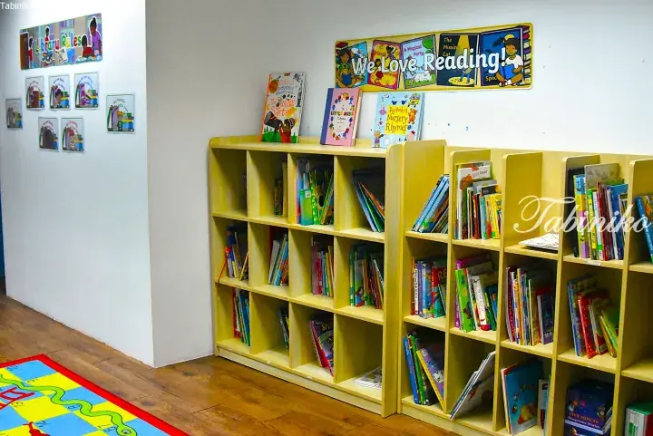 Light wood bookshelves filled with children&rsquo;s books in a bright library corner with reading posters.