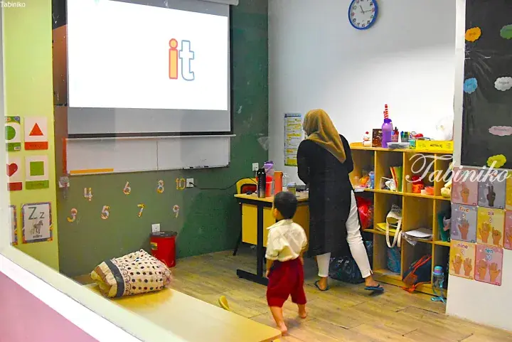 A young boy walks in a classroom while a teacher prepares materials near a projector screen displaying the letters &lsquo;it&rsquo;.