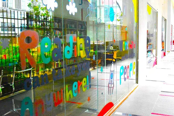 Colorful reading-themed window decals at a modern library or bookstore, featuring the quote &lsquo;Reading can take you anywhere!&rsquo; and playful rocket illustrations.