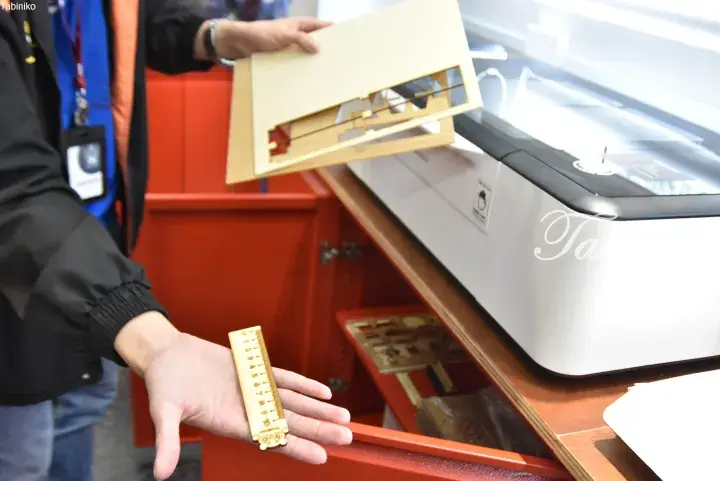 Person holding laser-cut wooden ruler and model pieces next to a Tabiniko laser engraving machine.