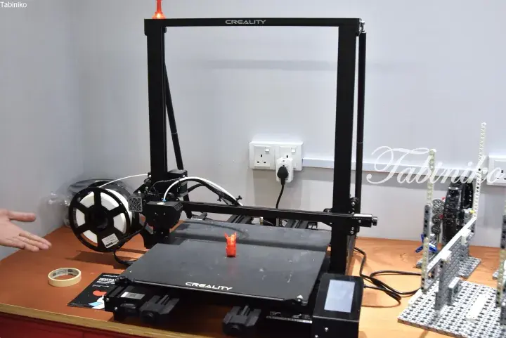 A black Creality 3D printer on a wooden desk with filament spools and a small orange printed figurine.