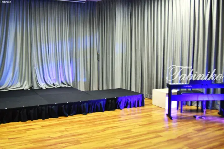 A small stage with black drapes and blue lighting in a hall with wooden flooring and a Casio keyboard.
