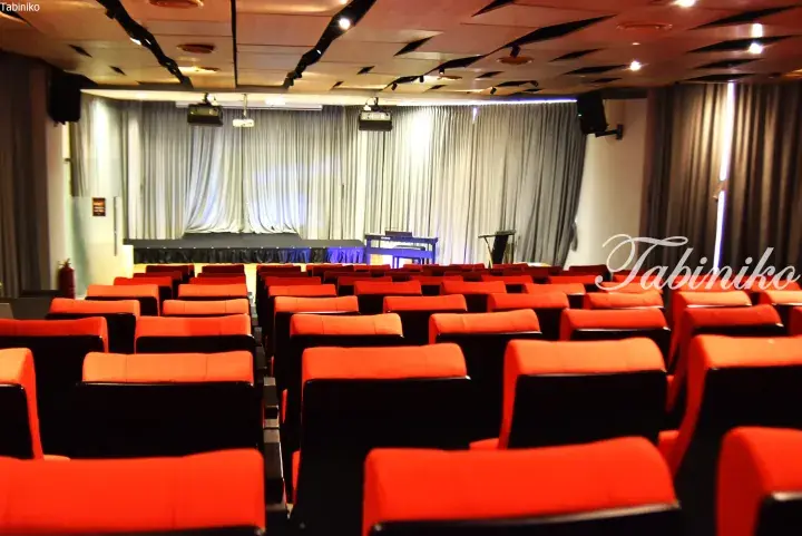 Rows of red and black seats in a modern auditorium with a stage, curtains, and ceiling lights.