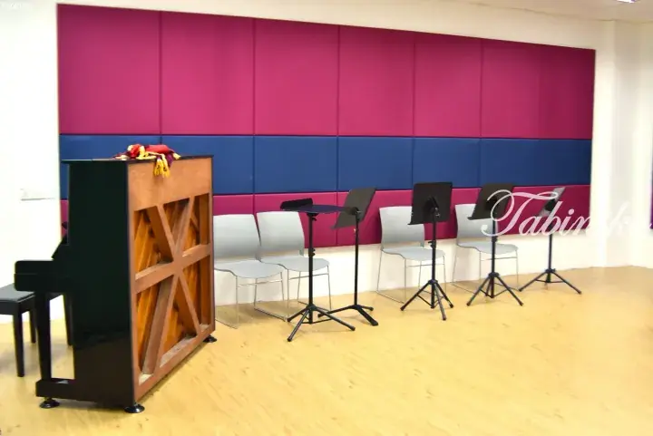 Music practice room with a piano, chairs, and music stands against a pink and blue acoustic wall.