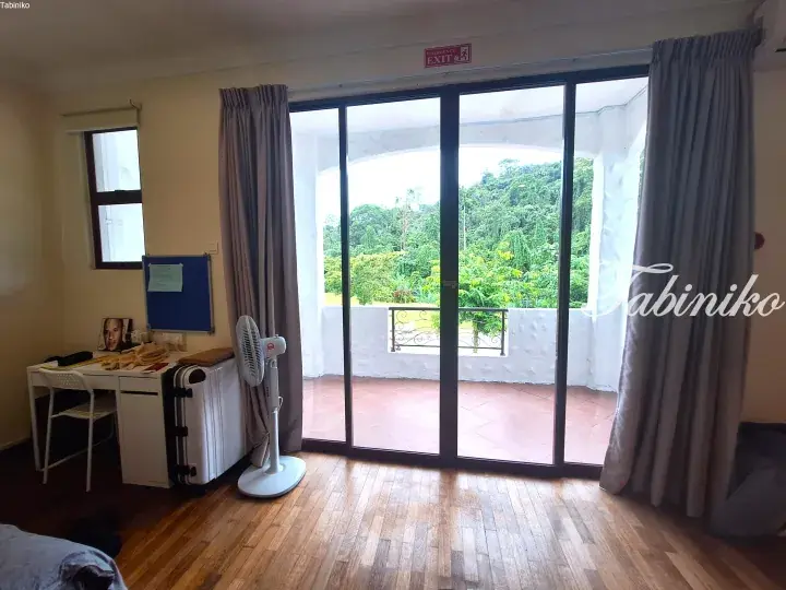 Bright bedroom with wooden floors and a balcony overlooking lush greenery, featuring a white desk and standing fan.