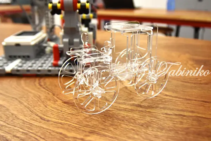 Transparent acrylic model of a vintage carriage on a wooden table with a Lego robot in the background.