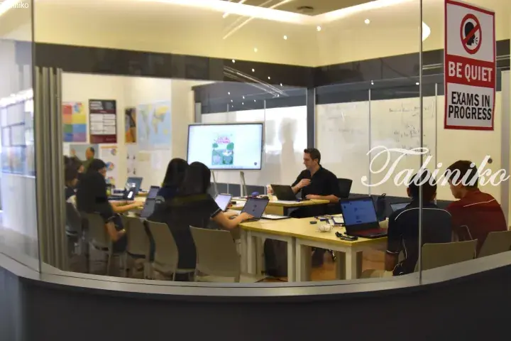 Students studying in a glass-walled room with a &lsquo;Be Quiet, Exams in Progress&rsquo; sign visible on the door.