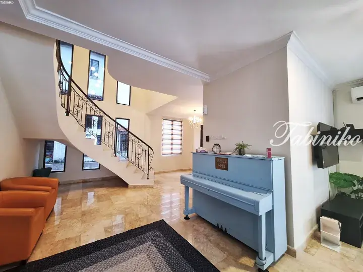 Bright living room with a curved staircase, blue piano, and orange sofa in a modern Malaysian home.