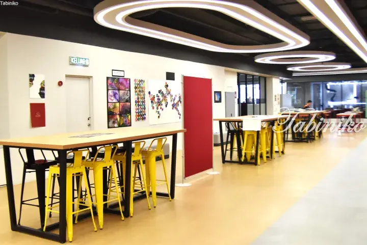 Modern co-working space with yellow stools, wooden tables, and art displays under unique ceiling lights.
