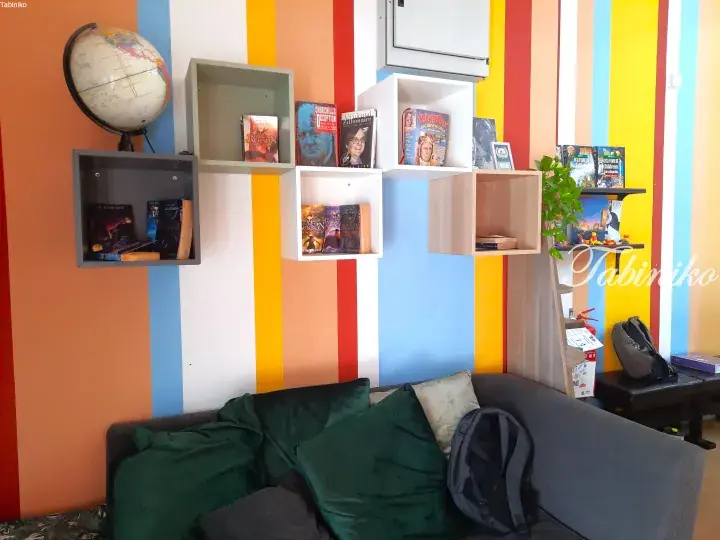 Colorful striped wall with floating shelves holding books, a globe, and a grey sofa with green cushions in a cozy reading nook.