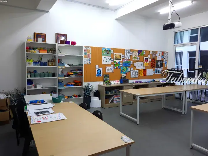 Bright art classroom with student artwork on corkboard, organized shelves, and long wooden tables for creative activities.