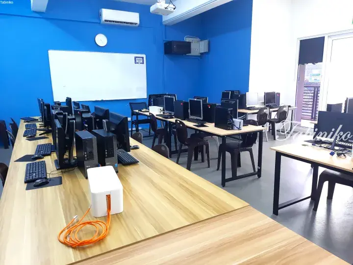 Modern computer lab with rows of desktop PCs on light wood desks against bright blue walls and a whiteboard.