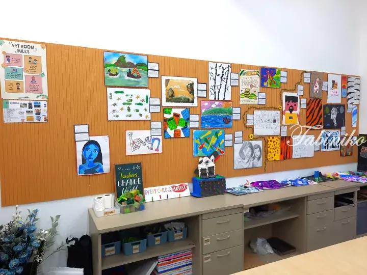 Colorful children&rsquo;s artwork displayed on a corkboard in a bright art room with organized storage cabinets below.