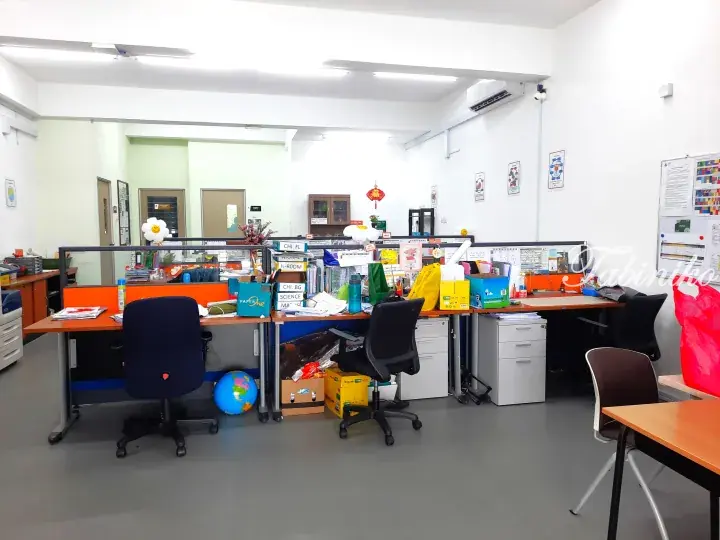 A bright office space with orange desks, black chairs, and a globe, likely a school staff room in Malaysia.