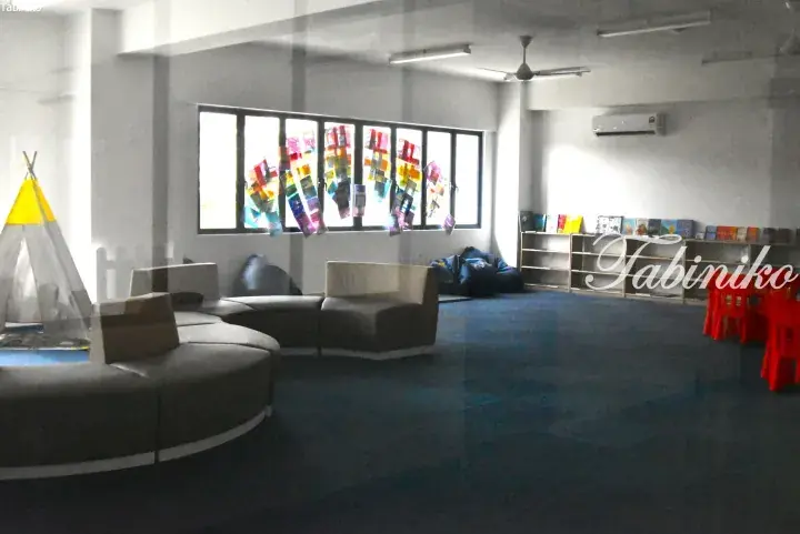 Modern library reading area with curved grey sofas, a teepee tent, bookshelves, and colorful artwork on windows.