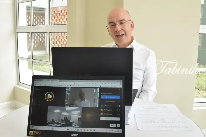 A man in a white shirt smiles during a Jitsi video call on an Acer laptop, with handwritten notes on the table.