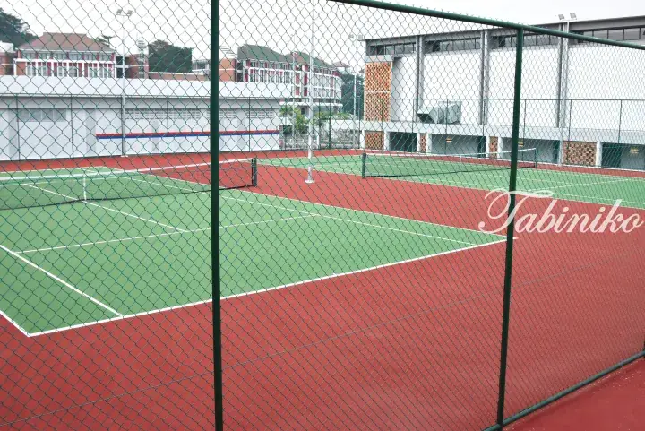 Red and green hard tennis courts viewed through a green chain-link fence with surrounding buildings in the background.