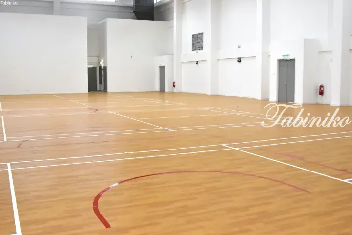 Spacious indoor sports hall with wooden flooring marked for badminton and basketball courts, featuring white walls and exit doors.