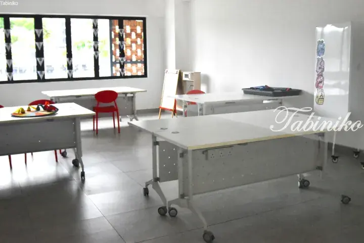 Bright classroom with white modular tables on wheels, red chairs, and large windows overlooking greenery.