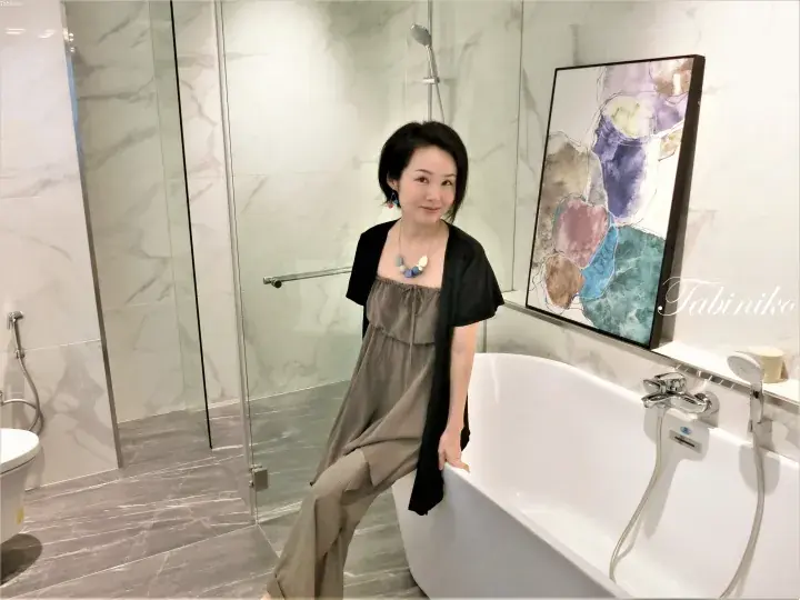 Woman in grey dress leaning on a white bathtub in a modern marble bathroom with glass shower and abstract art.