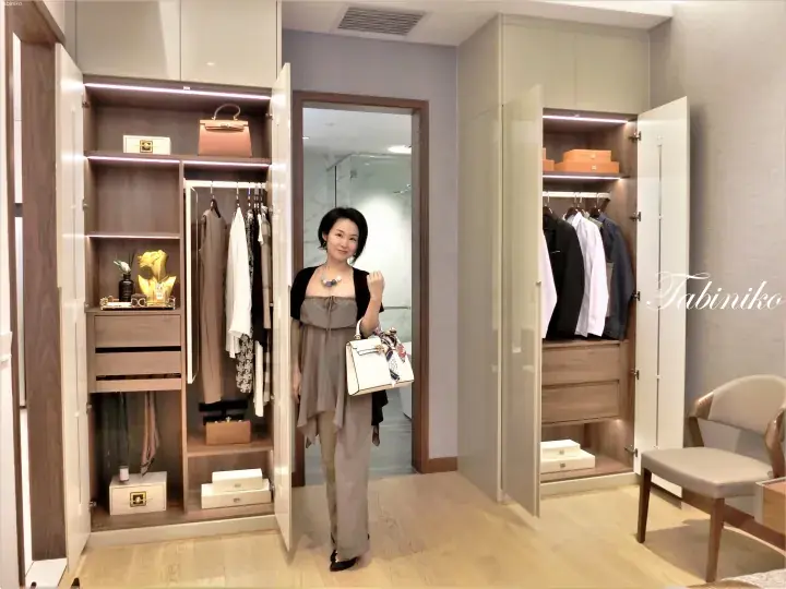 Woman standing in a modern walk-in closet with open wardrobes displaying luxury handbags and organized clothing.