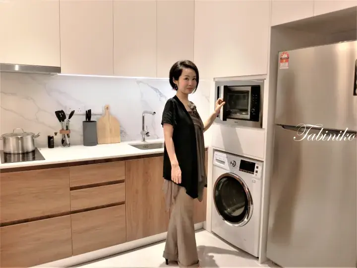 Woman standing in a modern kitchen with wooden cabinets, a microwave, and a washing machine.