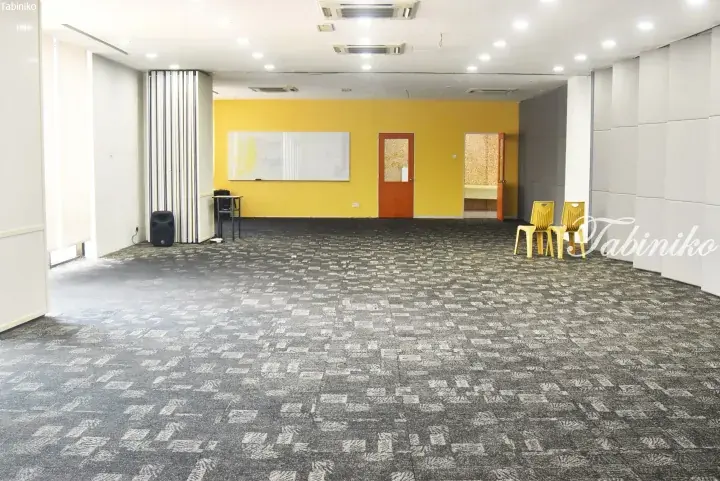 Spacious event hall with grey patterned carpet, yellow accent wall, whiteboard, and yellow chairs.
