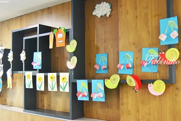 Colorful children’s crafts including snails and cherries displayed on a wooden wall with a black shelving unit.