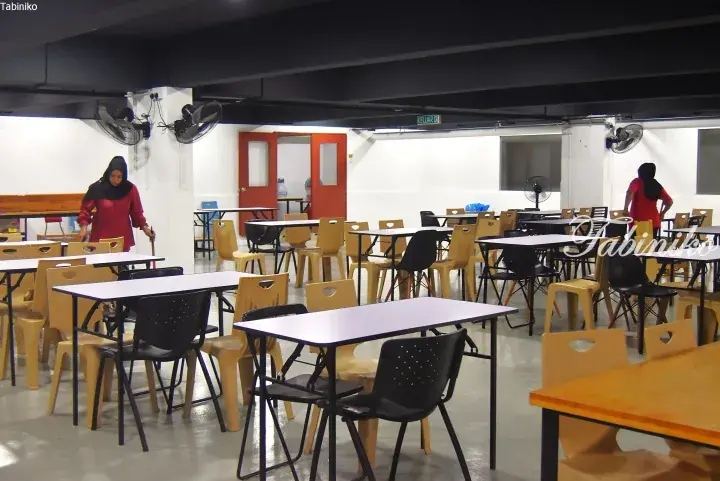 A spacious dining hall with white tables and beige chairs, featuring staff in red uniforms and a ‘KELUAR’ exit sign.