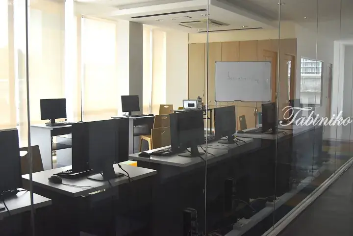Modern glass-walled office space with rows of computer workstations and a whiteboard.