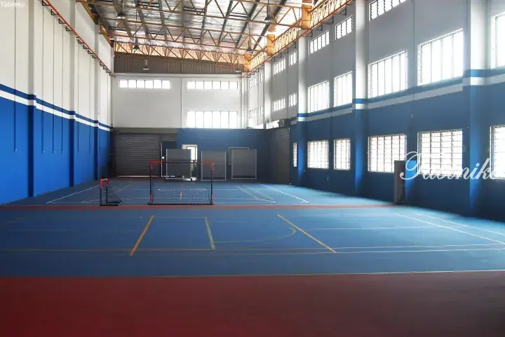 Spacious indoor sports hall with blue flooring, high ceilings, and large windows for natural light.
