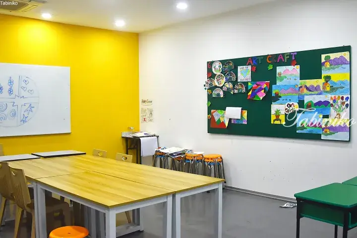 Bright classroom with yellow walls, wooden tables, and a green bulletin board displaying children’s art and craft projects.