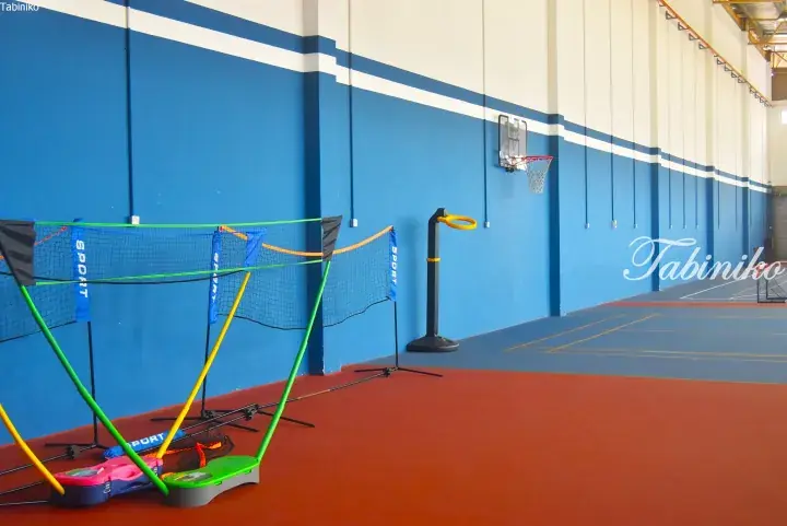 Colorful portable badminton nets and a basketball hoop in a spacious indoor sports hall with blue and white walls.