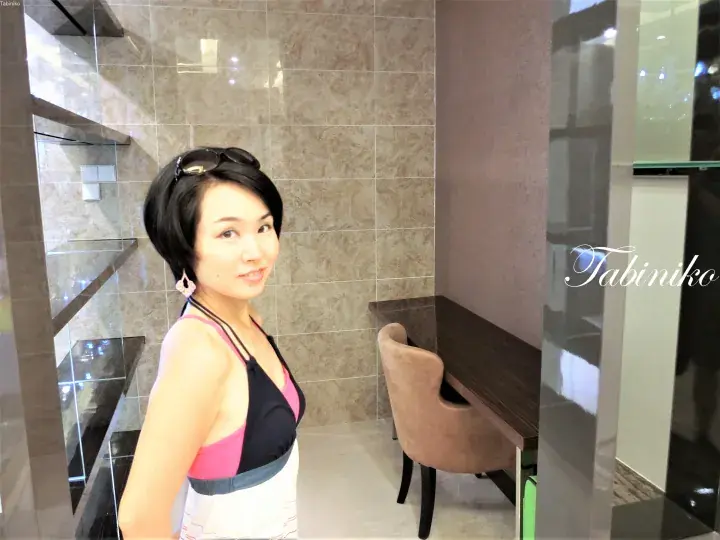 水着を着た女性が、ベージュの大理石タイルと洗面台が備わったモダンなバスルームでポーズを取る。