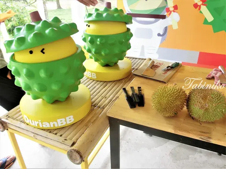 Cute durian-themed mascot statues on a bamboo table next to real durians and tools at DurianBB.